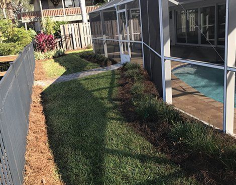Lawn with a gray fence, paving stones, and plants next to a screened pool enclosure and building.