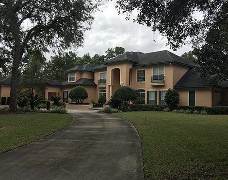 Large tan house with a long driveway, on a grassy lawn under a cloudy sky.