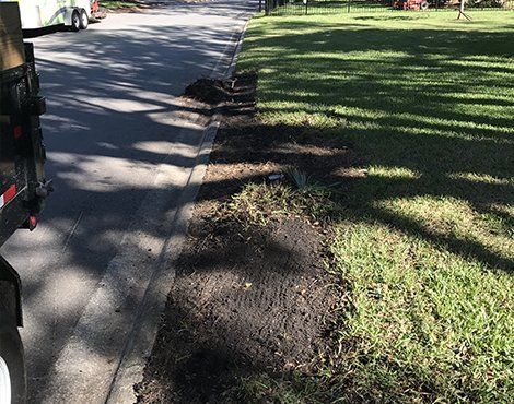 A curb with freshly turned soil borders grass and a road, trailer parked on the left.