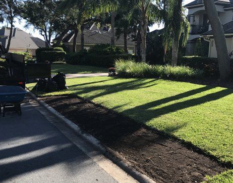 A freshly dug garden bed bordering a sidewalk and lawn, with dark soil, wheelbarrow, and trailer.