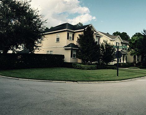 Two-story beige house on a corner lot with a well-manicured lawn and trees under a cloudy sky.