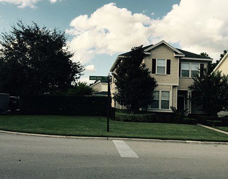 Two-story beige house on corner lot with green lawn, trees, and street sign under a cloudy sky.