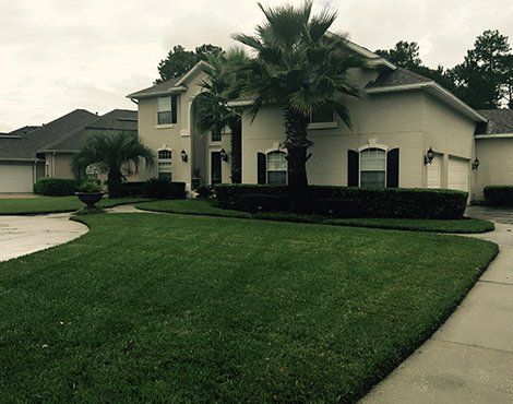 Two-story beige house with lush green lawn and palm trees. Cloudy day.