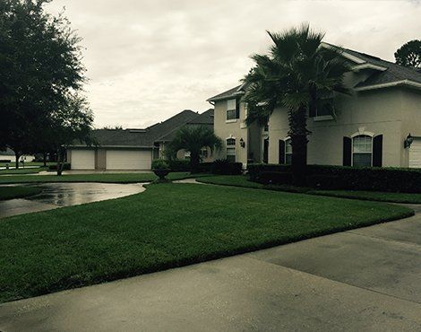 Large house with well-manicured green lawn, palm trees, and a cloudy sky. A driveway is in the foreground.