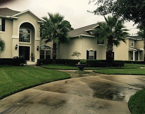 Large beige house with arched entry, palm trees, and manicured lawn. Wet driveway.