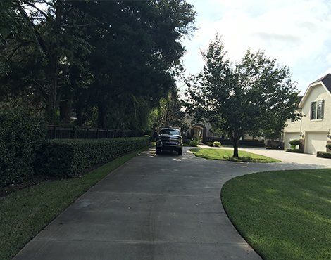 A black SUV parked on a concrete driveway leading to a house. Green lawn and trees surround the driveway.