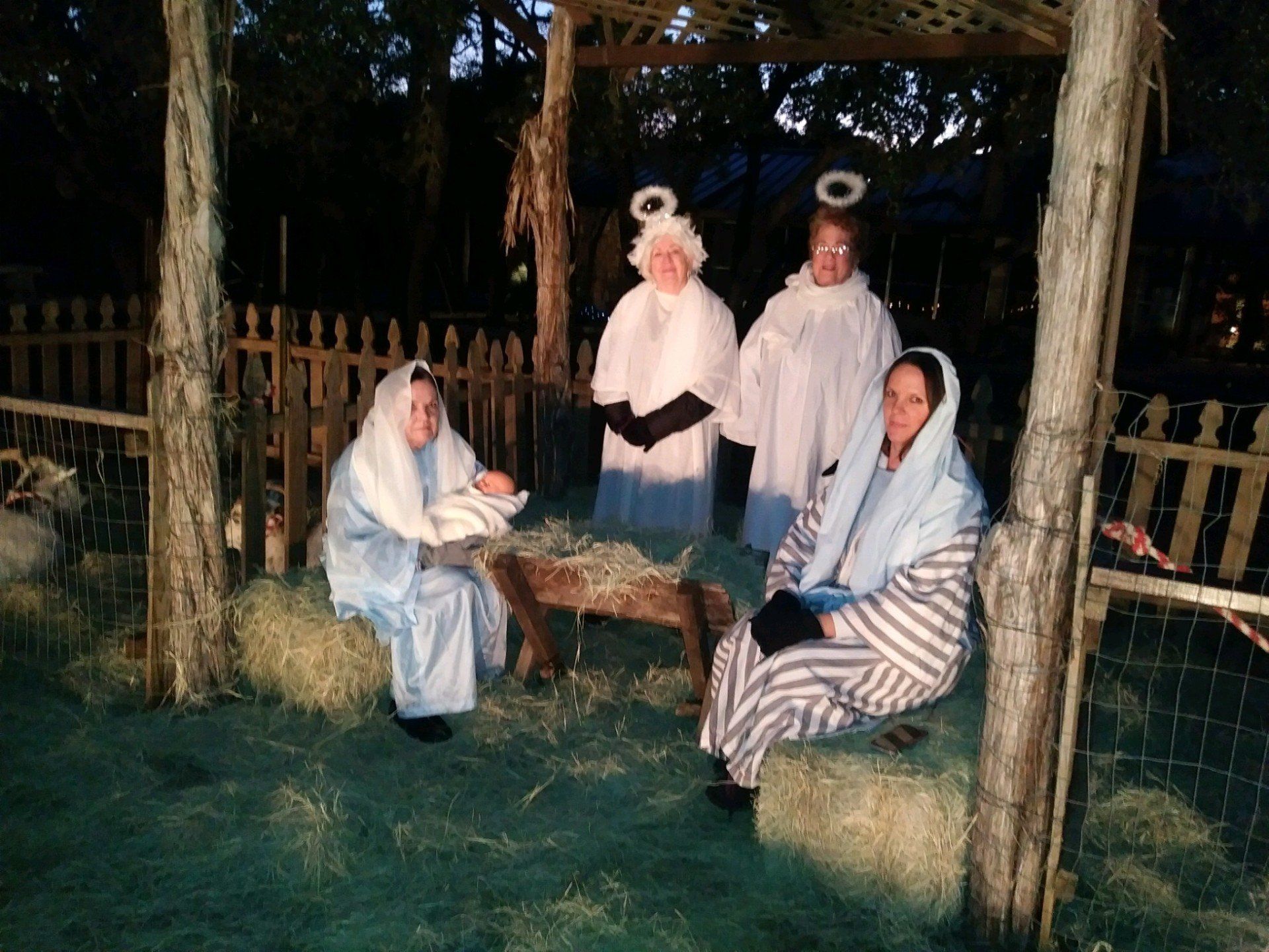 Nativity scene: people dressed as Mary, Joseph, angels; manger with hay; outdoor setting.