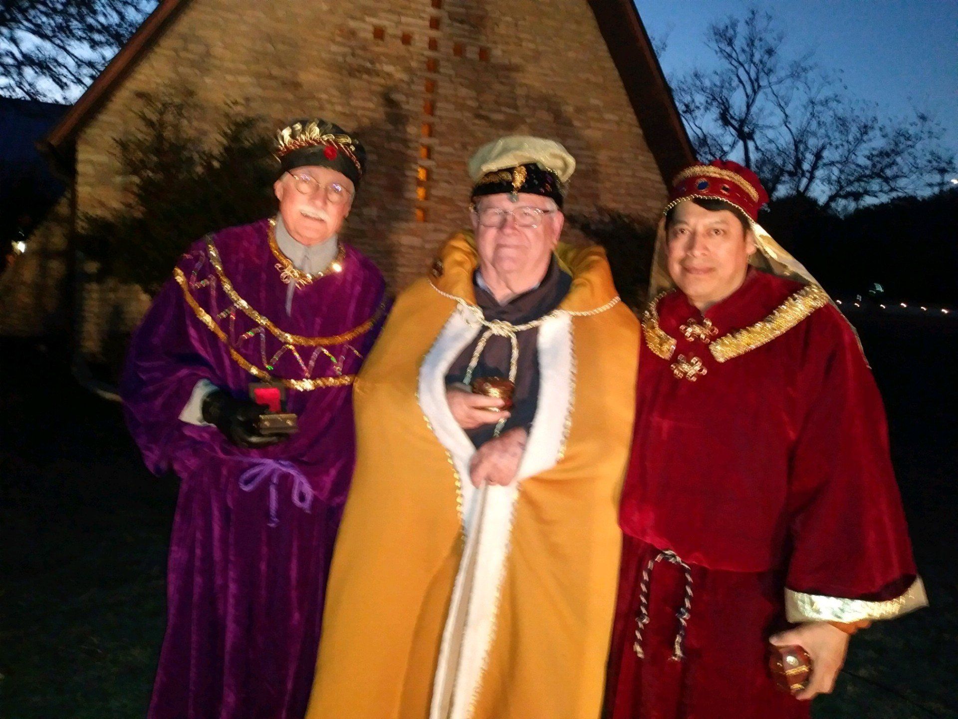 Three people in colorful robes holding small cups, standing outdoors in front of a building at dusk.