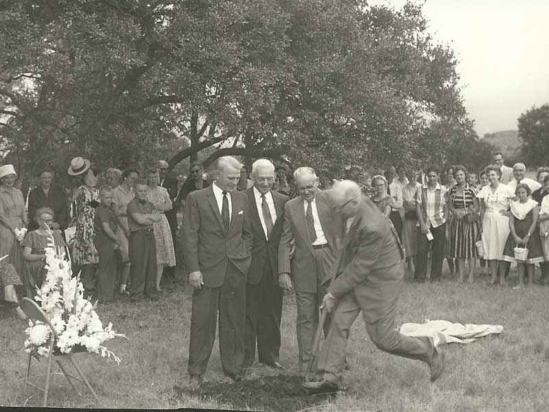 Men in suits breaking ground with a shovel, surrounded by a crowd outdoors.