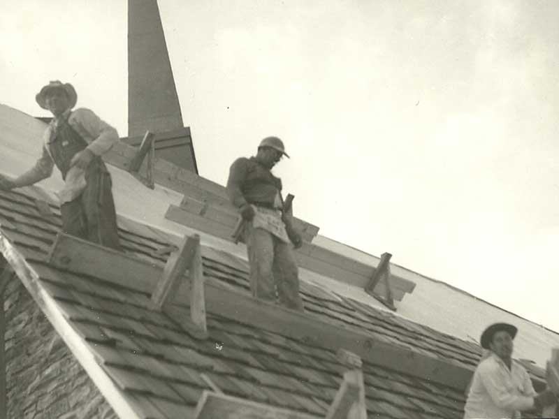 Three roofers working on a sloped roof.  They are using wood supports.. A church steeple is in the background.