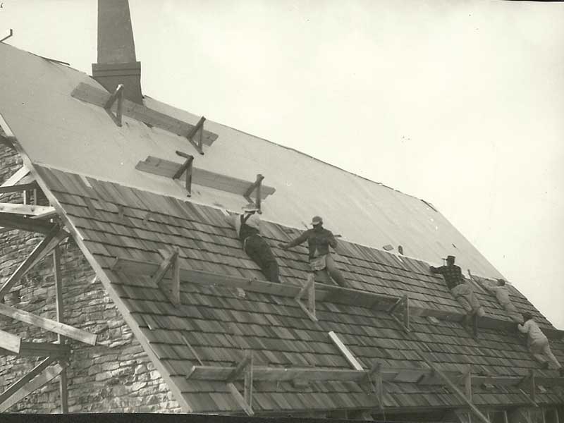 Men working on a roof, installing shingles. The partially completed roof has exposed paper and wooden scaffolding.