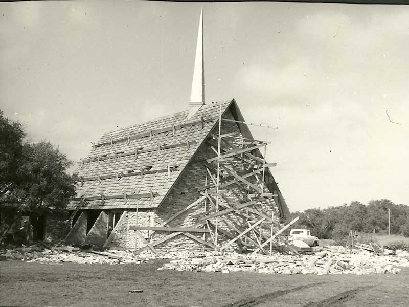 Church under construction, A-frame design, wooden scaffolding, stone walls, tall spire, rural setting.