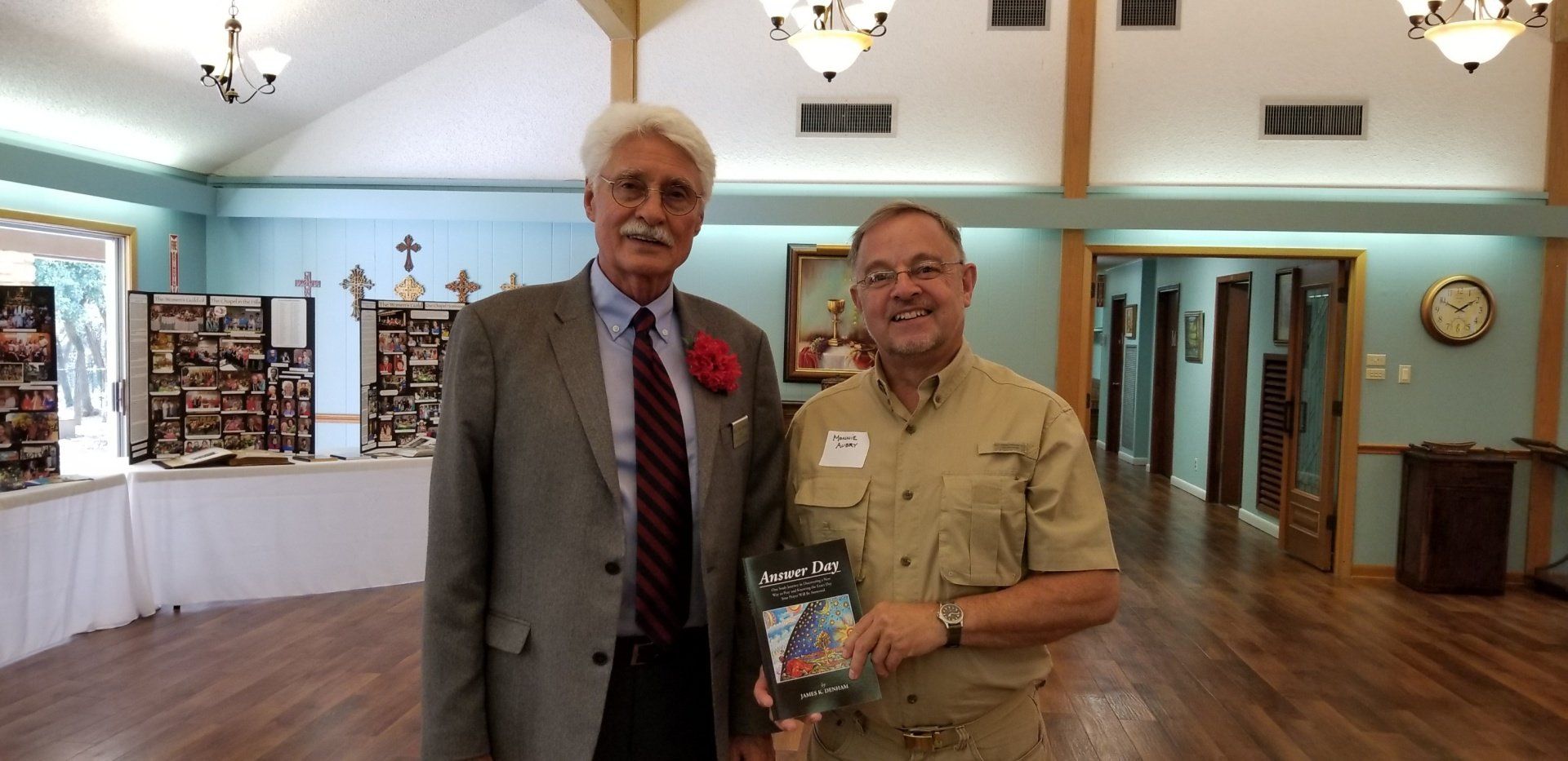 Two men in a light-filled room pose for a photo. One holds a book. The setting appears to be a reception or event.