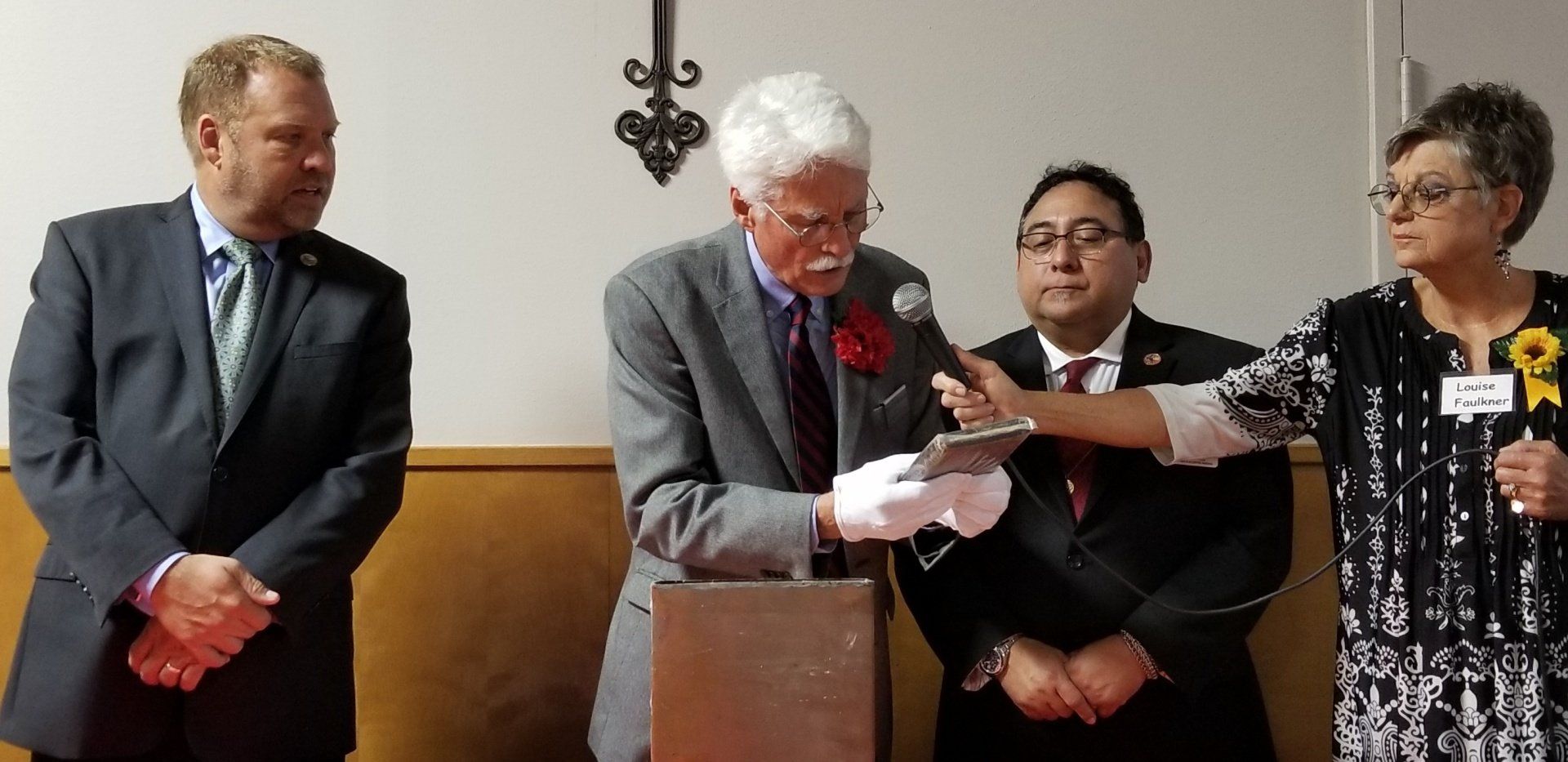 Four people at a podium. A man in a suit reads from a plaque.  A cross hangs in the background.