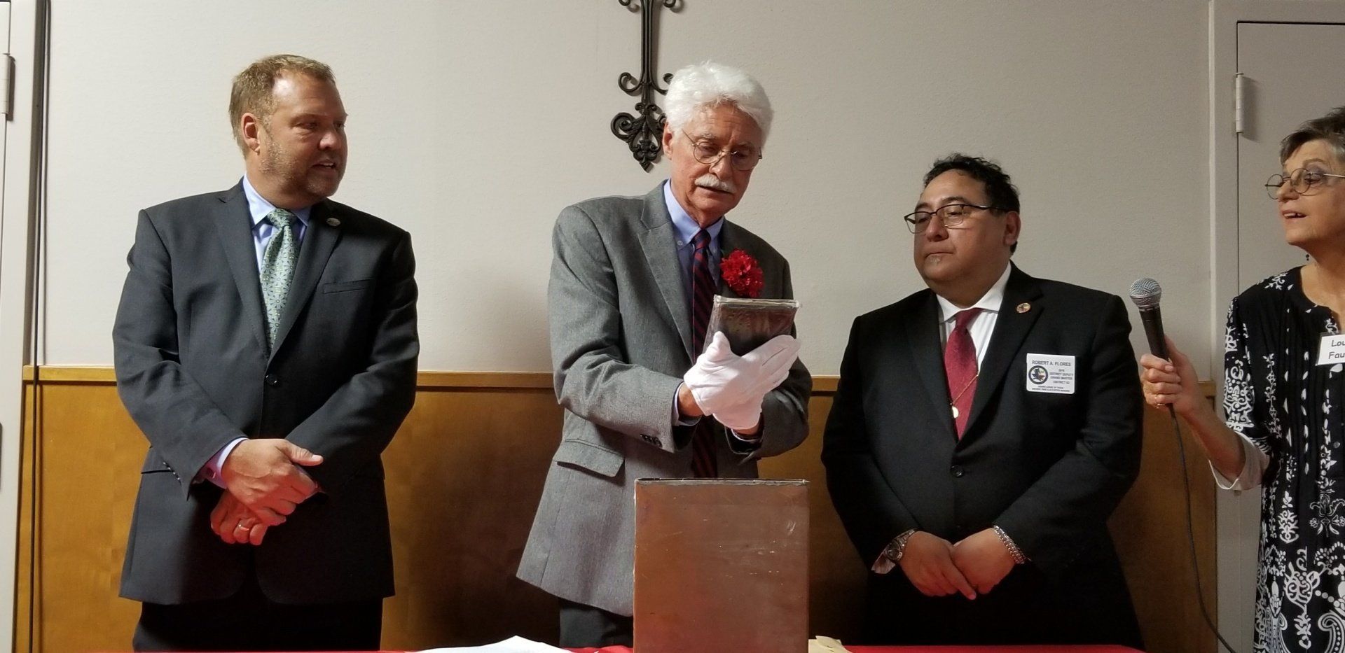 Four people at a presentation. A man in a suit holds a book wearing white gloves. Others in suits stand nearby.