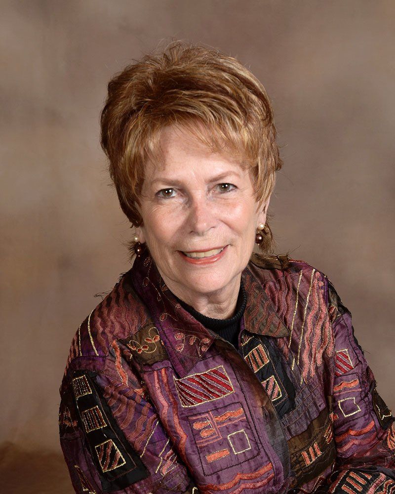 Woman with short auburn hair smiles, wearing a purple and black patterned jacket against a brown background.