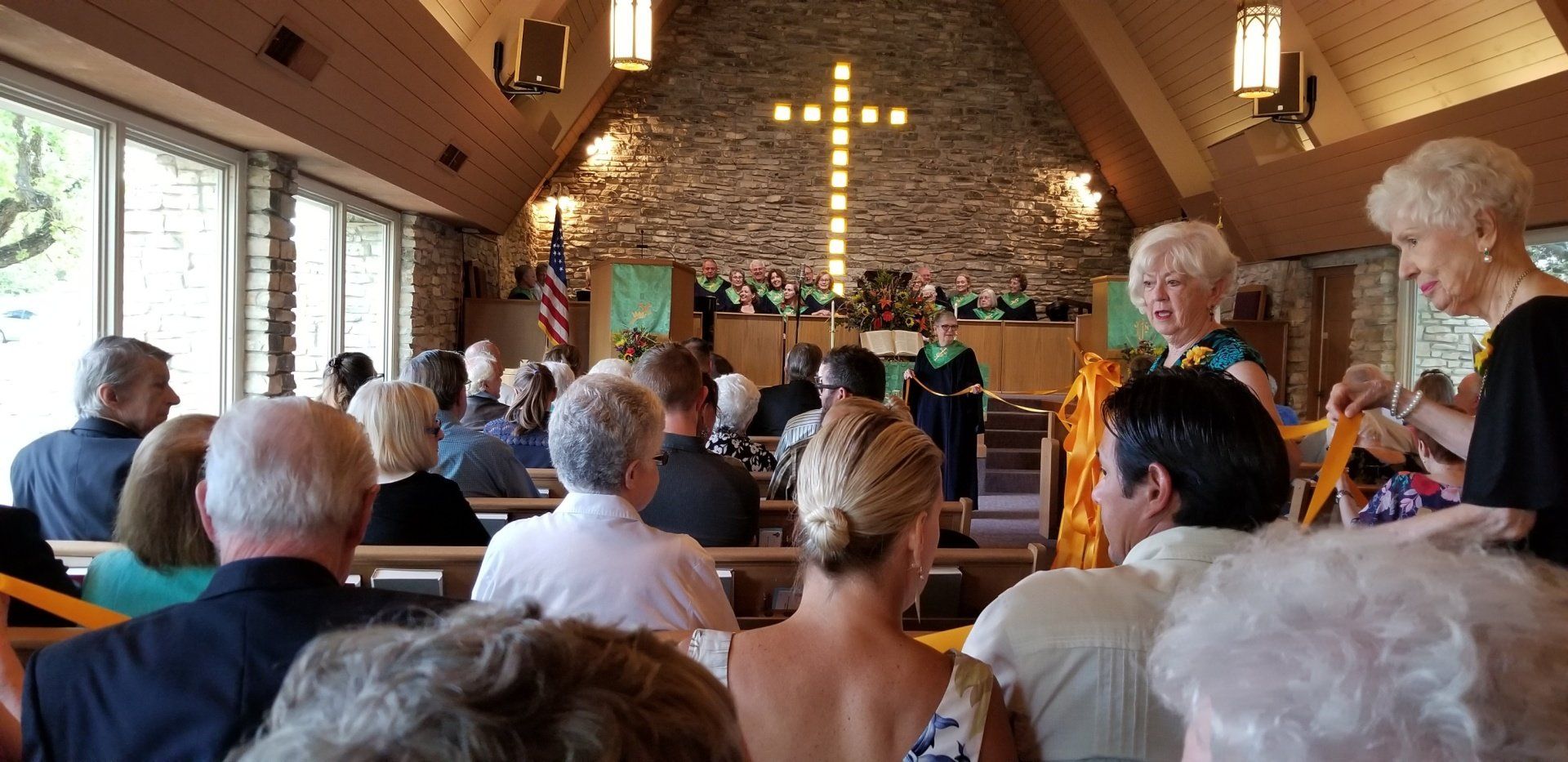 People attending a ceremony inside a church with a large cross on a stone wall.