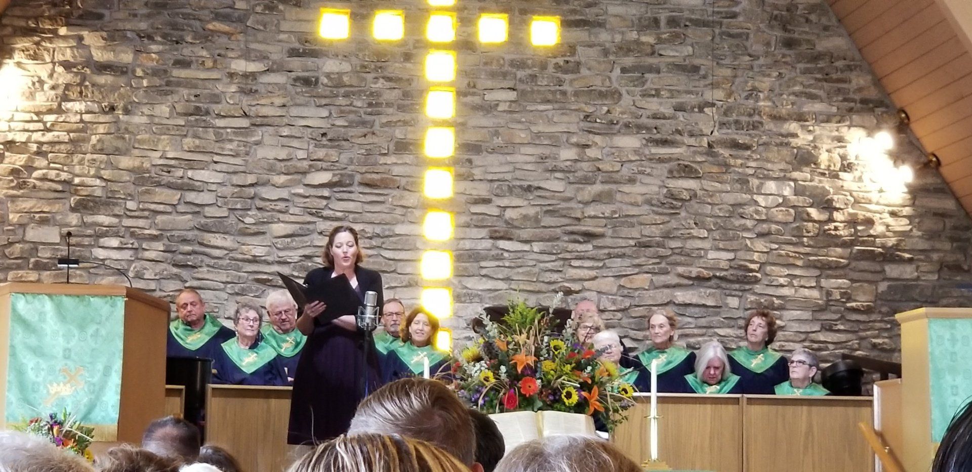 Woman speaking from a podium at a church with a choir and a glowing cross in the background.