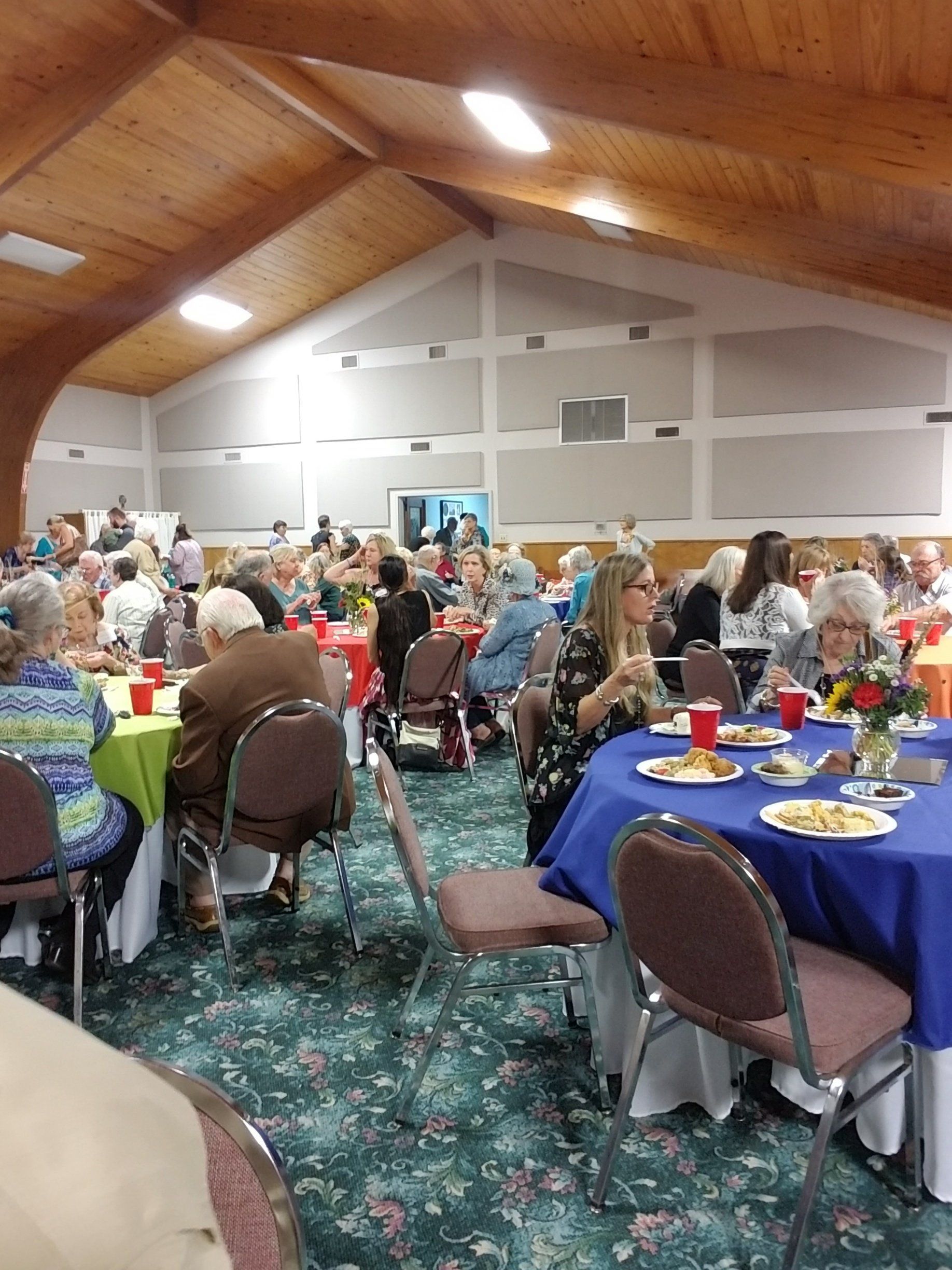 People seated at tables with colorful tablecloths in a large hall, likely an event or gathering.