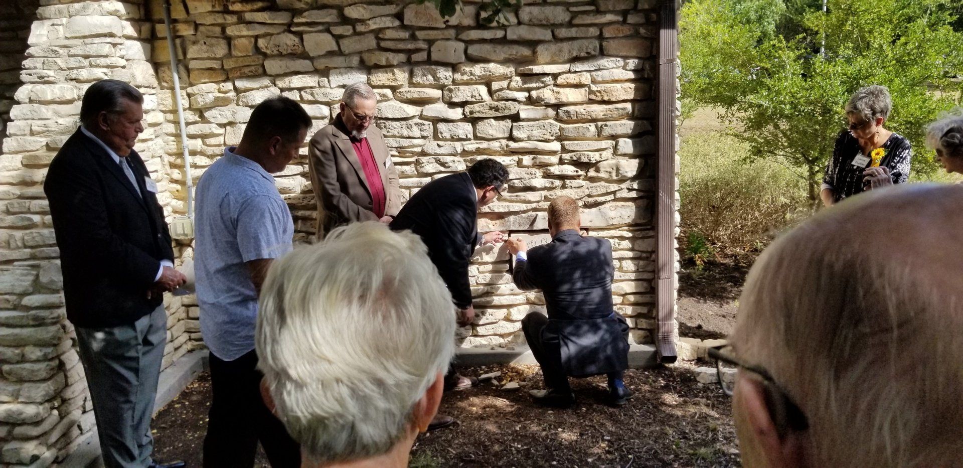 People gather around a stone wall, one kneeling and touching it. Outdoors, trees in the background.