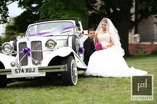Spirit Wedding Cars | A bride and groom are posing for a picture in front of a wedding car.