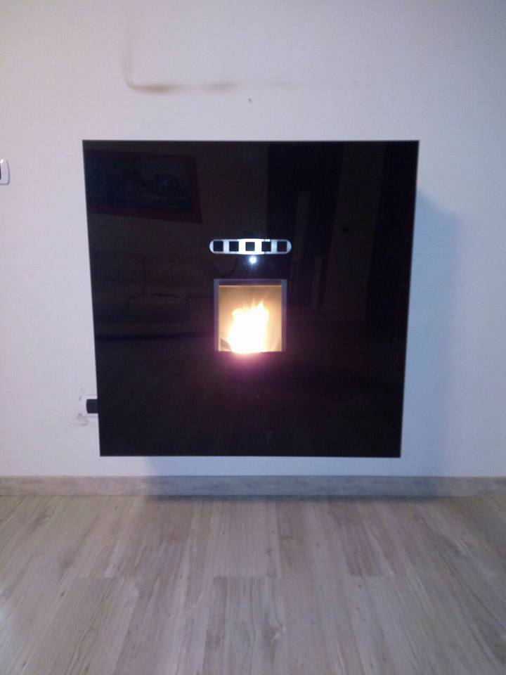 Caminetto nero a parete con fuoco scoppiettante in una stanza bianca, sopra un pavimento in legno chiaro.