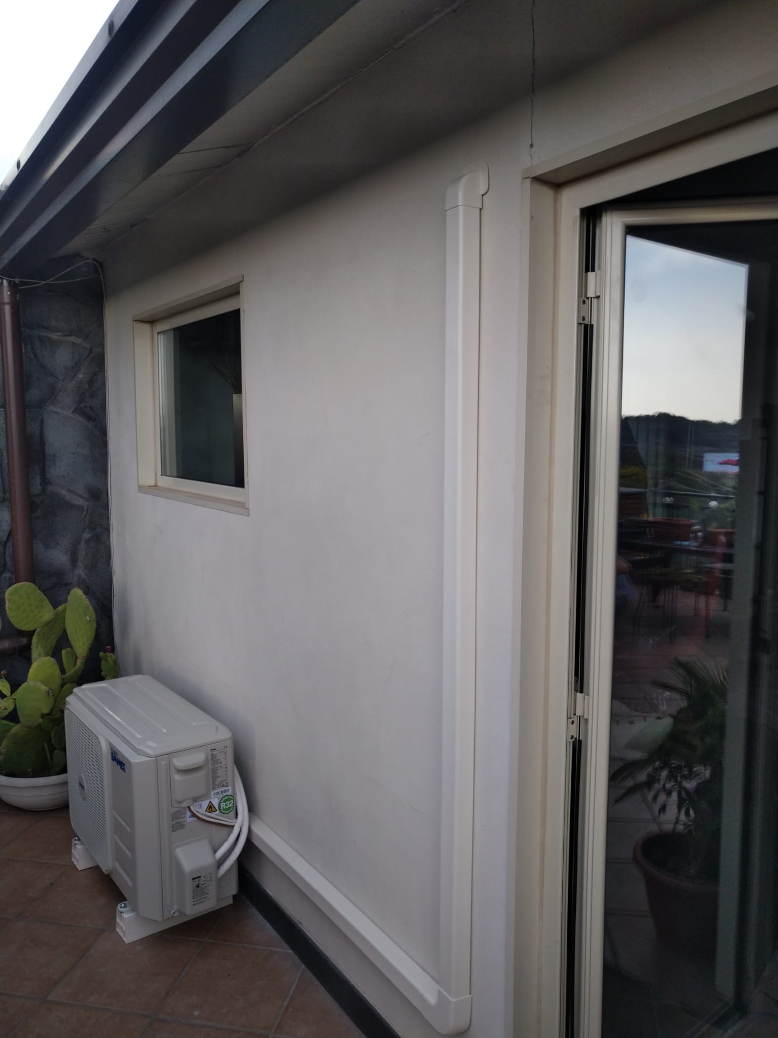 Parete esterna con finestra, porta scorrevole in vetro, condizionatore e piante di cactus su un patio.