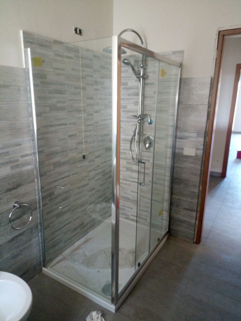 Bagno con box doccia in vetro, pareti rivestite con piastrelle grigie e porta con cornice in legno.