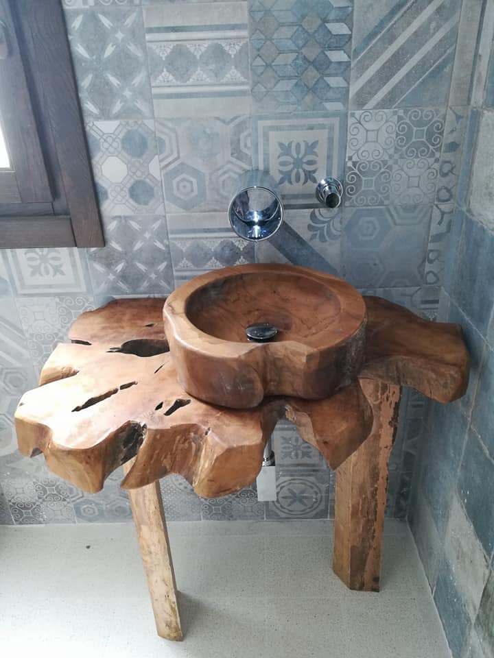 Lavabo rustico in legno su un supporto a forma di tronco, appoggiato a una parete rivestita di piastrelle decorate, con rubinetto cromato.