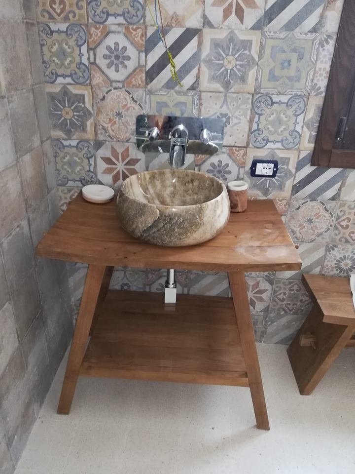 Mobiletto da bagno con lavabo in legno, piano in pietra e rubinetto cromato, addossato a una parete rivestita di piastrelle decorate.