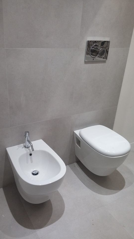 Bagno moderno con WC sospeso e piccolo bidet bianco su pavimento piastrellato grigio chiaro.