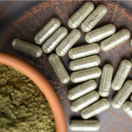 White capsules on a textured brown surface beside a bowl of green herb powder