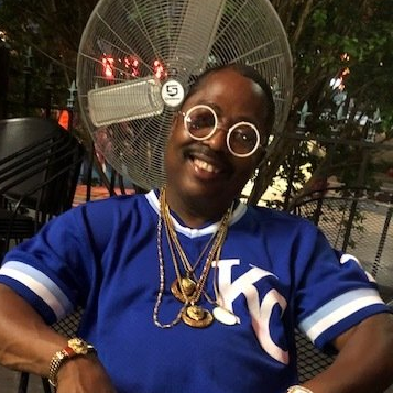 Smiling person in a blue KC jersey and round glasses sitting outdoors in front of a fan