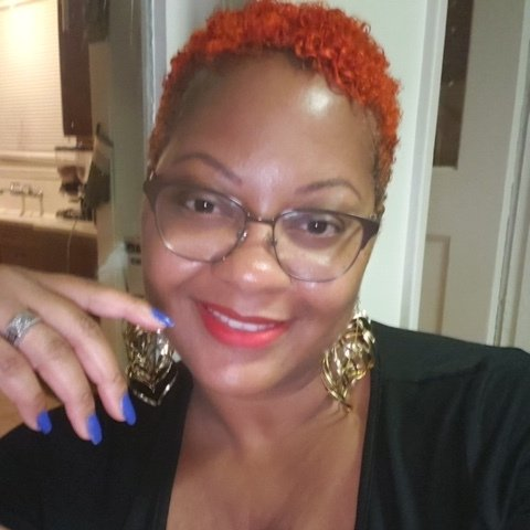 Smiling person with short red hair, glasses, and gold earrings, resting a hand near the face.