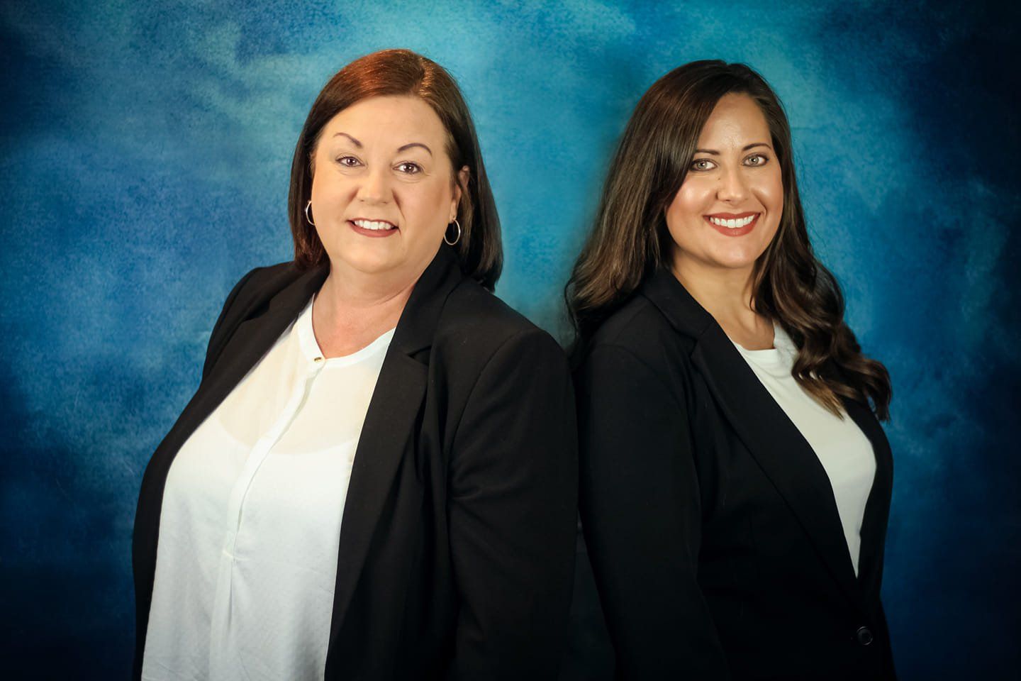 Two women in black blazers and white tops pose against a blue studio backdrop.