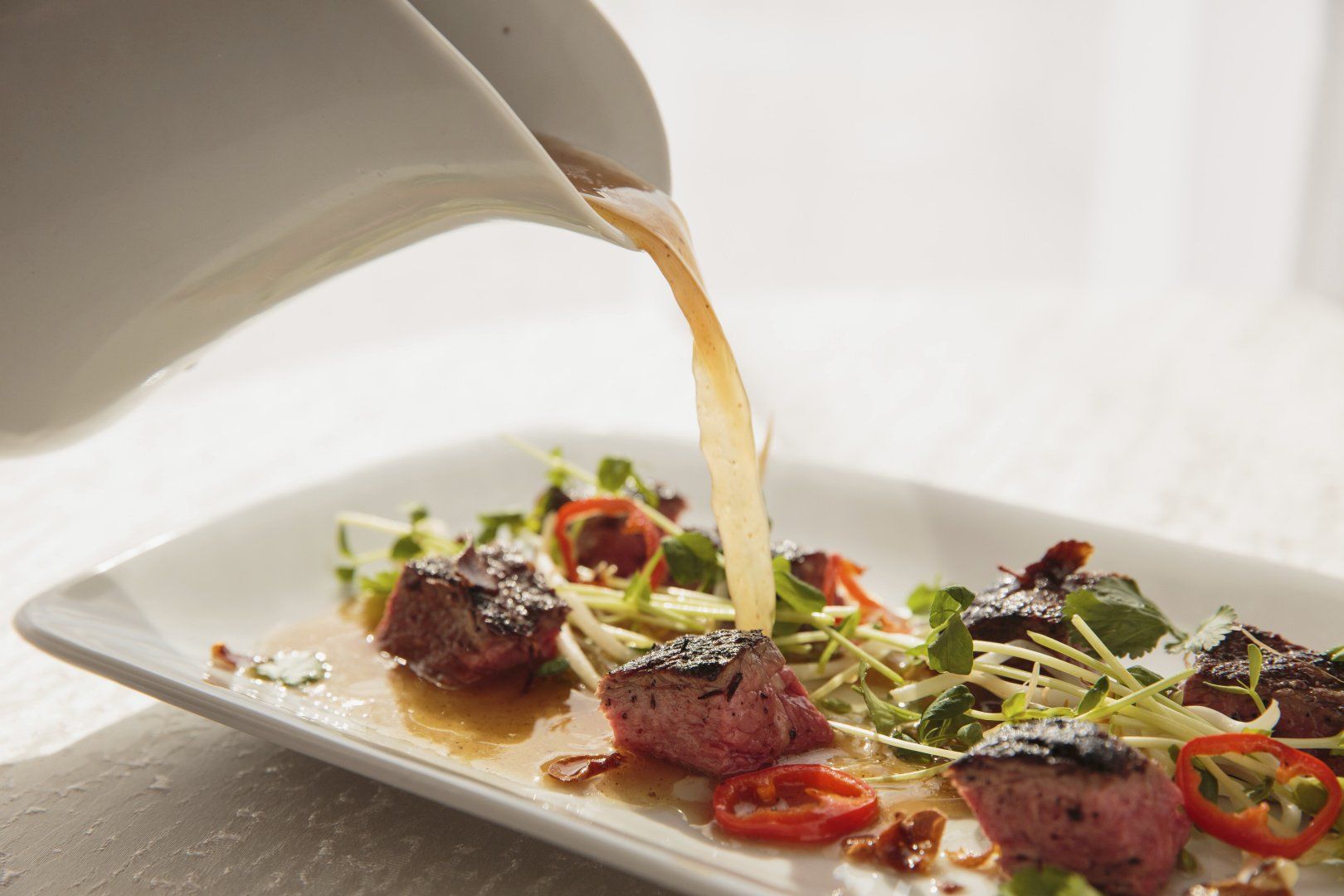 A white plate topped with meat and vegetables is being poured with sauce.