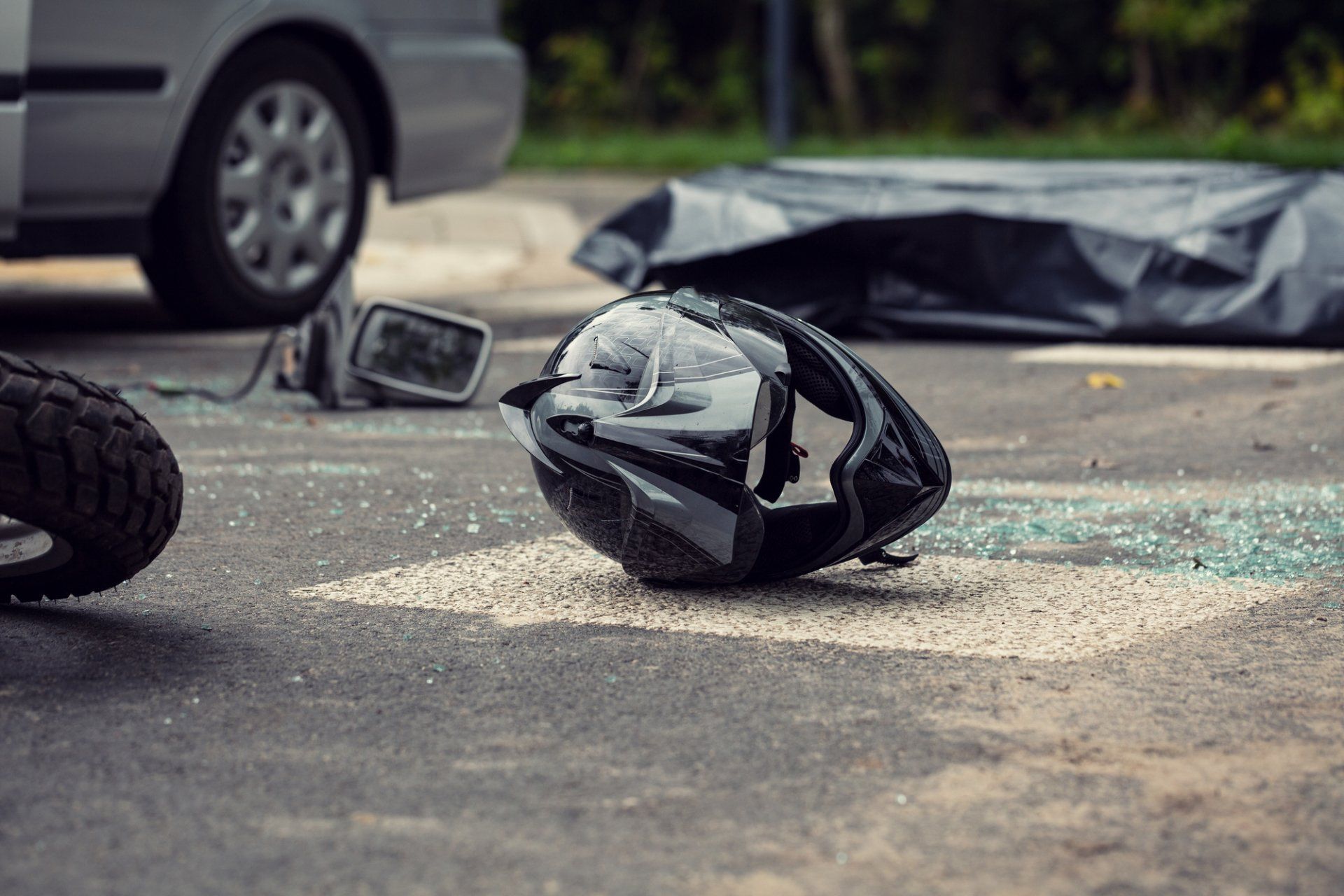 Motorcycle helmet damaged on pavement after crash; covered body and car visible in background.