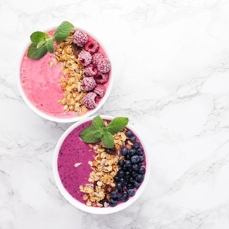 Two smoothie bowls: pink with raspberries, and purple with blueberries, granola, mint leaves.