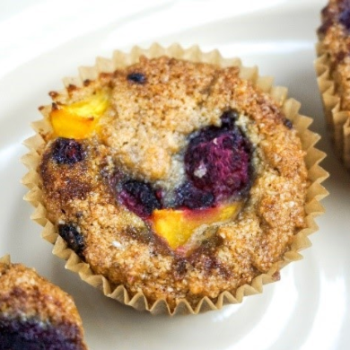 Muffin with visible berries and peach pieces in a paper liner on a white plate.