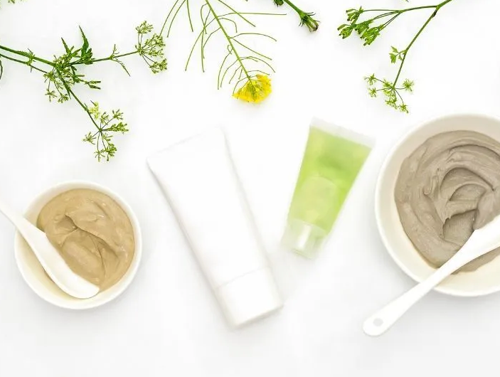 Skincare products: tubes and bowls of cream with sprigs of greenery on a white background.
