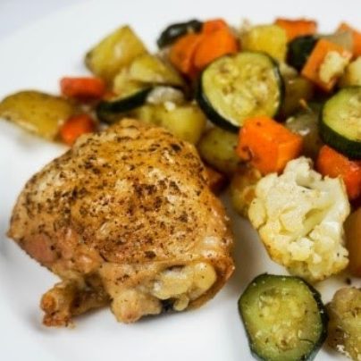 Roasted chicken thigh on a white plate with roasted vegetables, including zucchini and carrots.