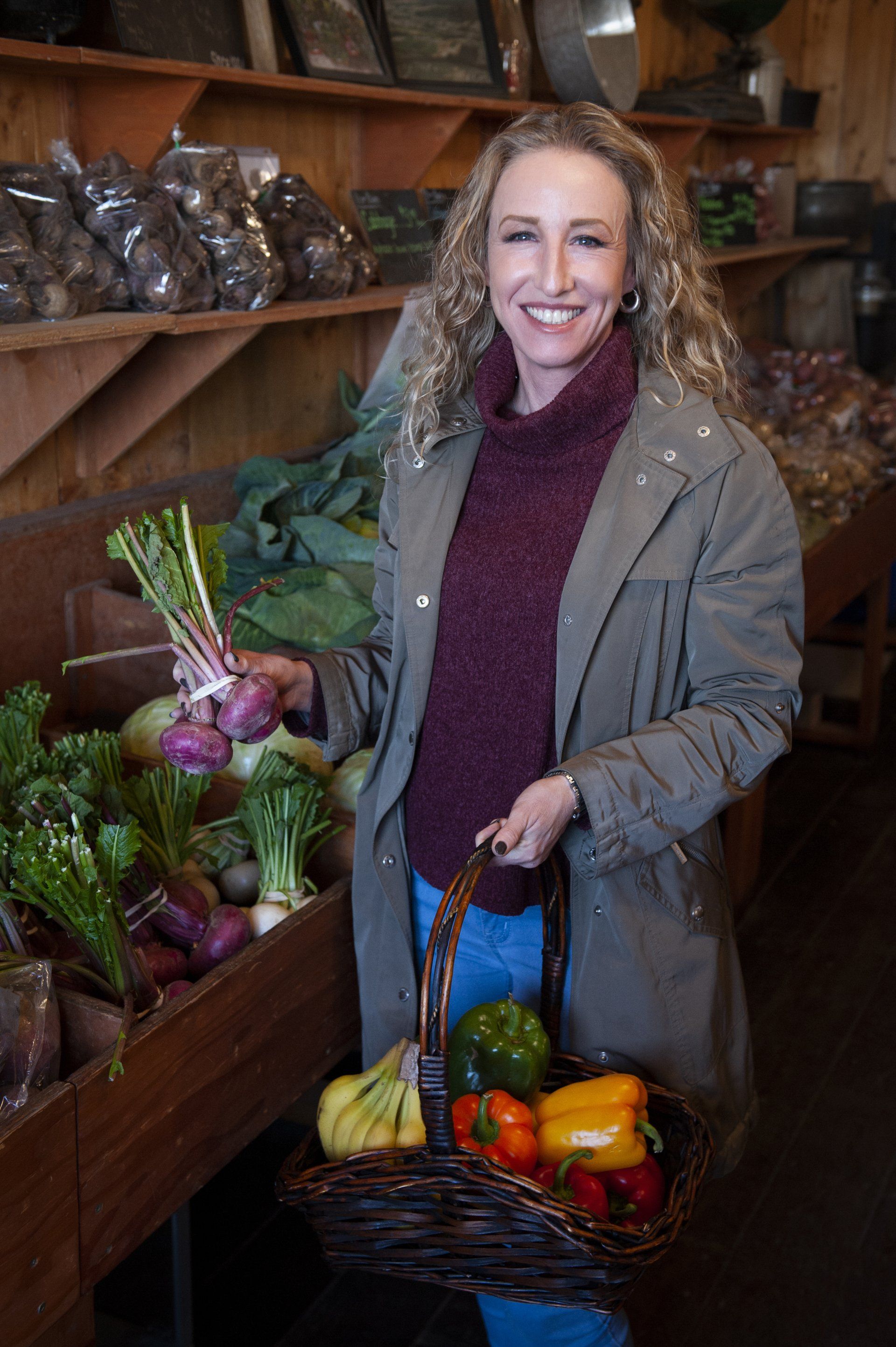Woman with curly hair smiles, holding basket of produce, armful of radishes at a farm stand.