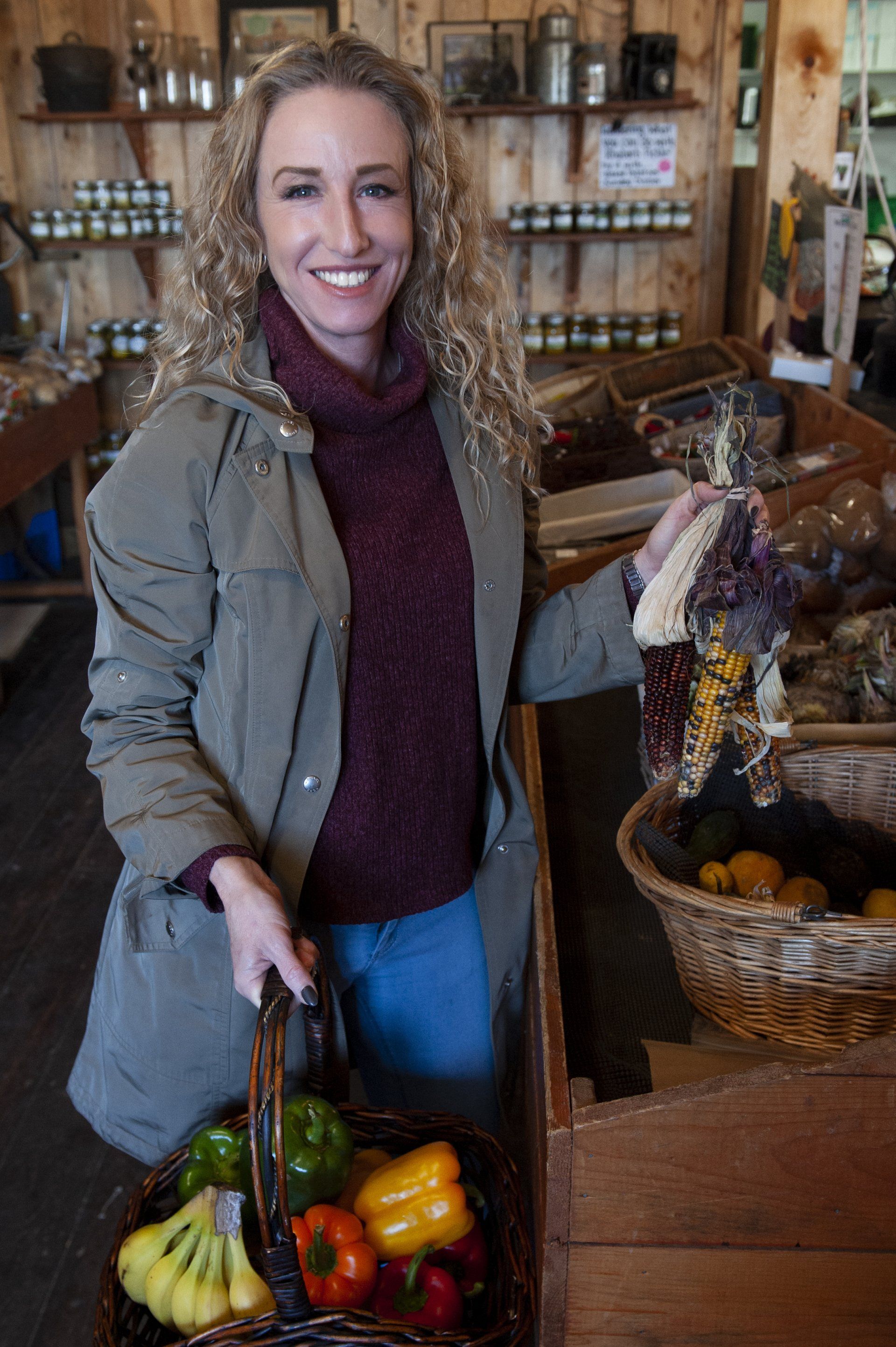 Woman with curly hair in a market, holding a basket of vegetables, smiling.