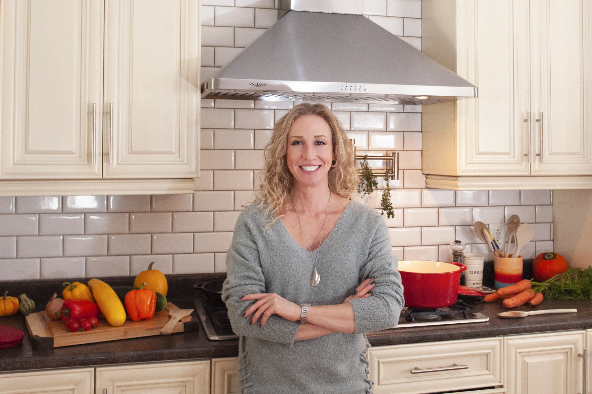 Woman in kitchen, arms crossed, smiling. Vegetables on counter, pot on stove, white cabinets.