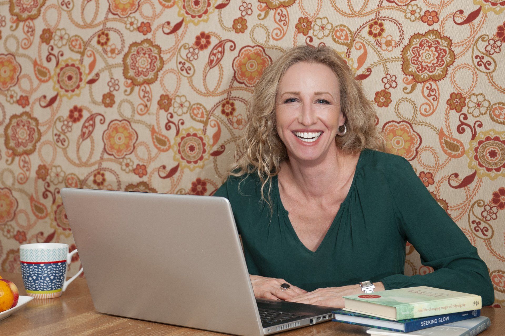Woman with laptop, smiling at desk with coffee mug and books, patterned wallpaper background.