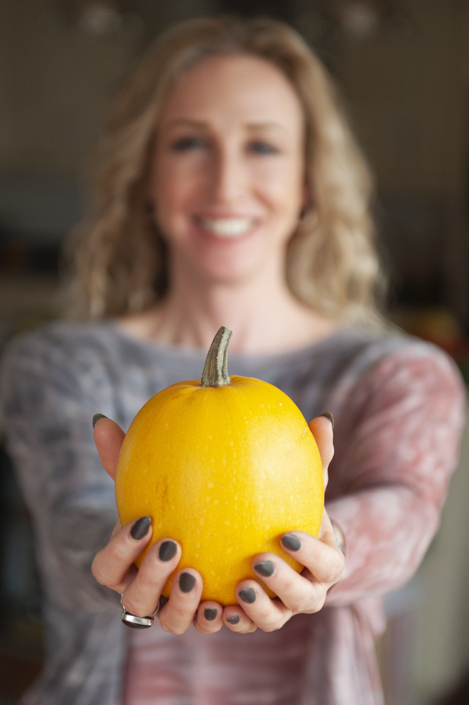 Woman smiling, holding out a small, yellow pumpkin.