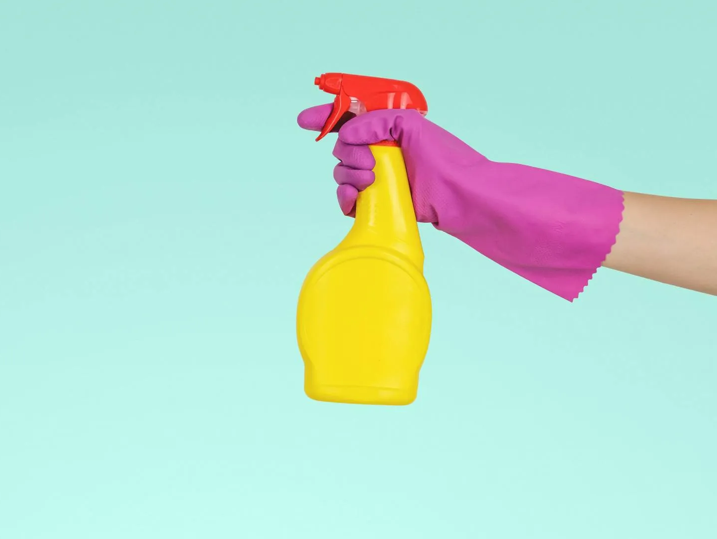 Hand in pink glove holding yellow spray bottle with red trigger, against a turquoise background.