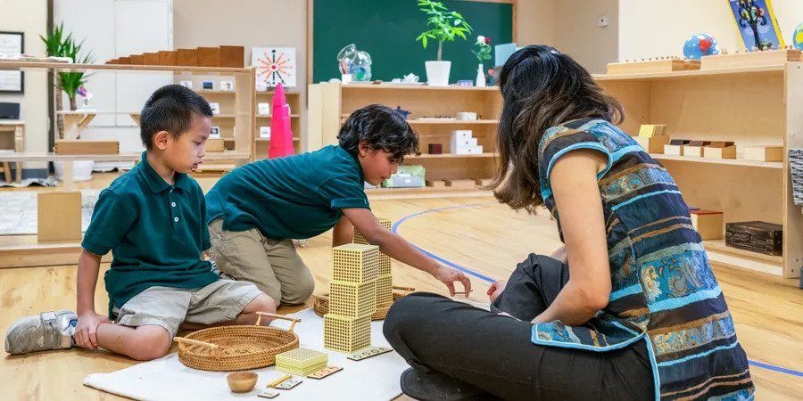 A montessori guide is sitting on the floor with two children learing with montessori math blocks.