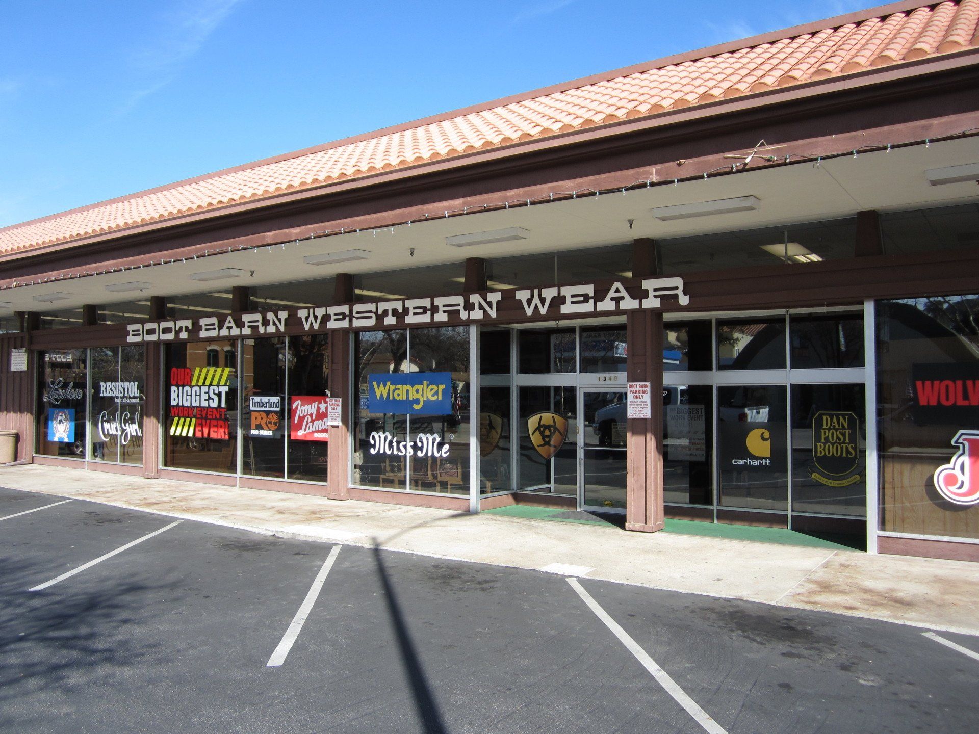 Western wear storefront in a strip mall with glass doors, sale signs, and a red tiled roof.