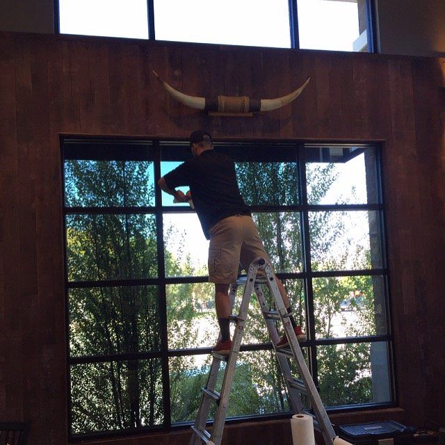 Person standing on a ladder adjusting a wall-mounted decor in a wood-paneled room with large windows.